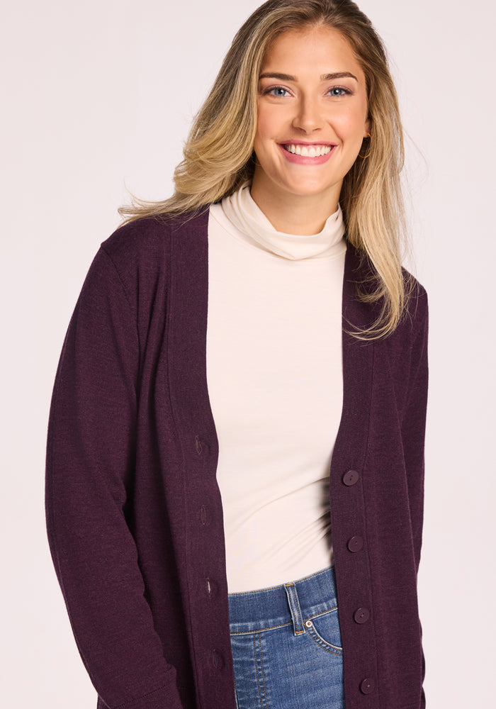 A smiling woman with long blonde hair wears the Woolx Gia Sweater in Deep Plum, paired with blue jeans, as she stands against a light background.
