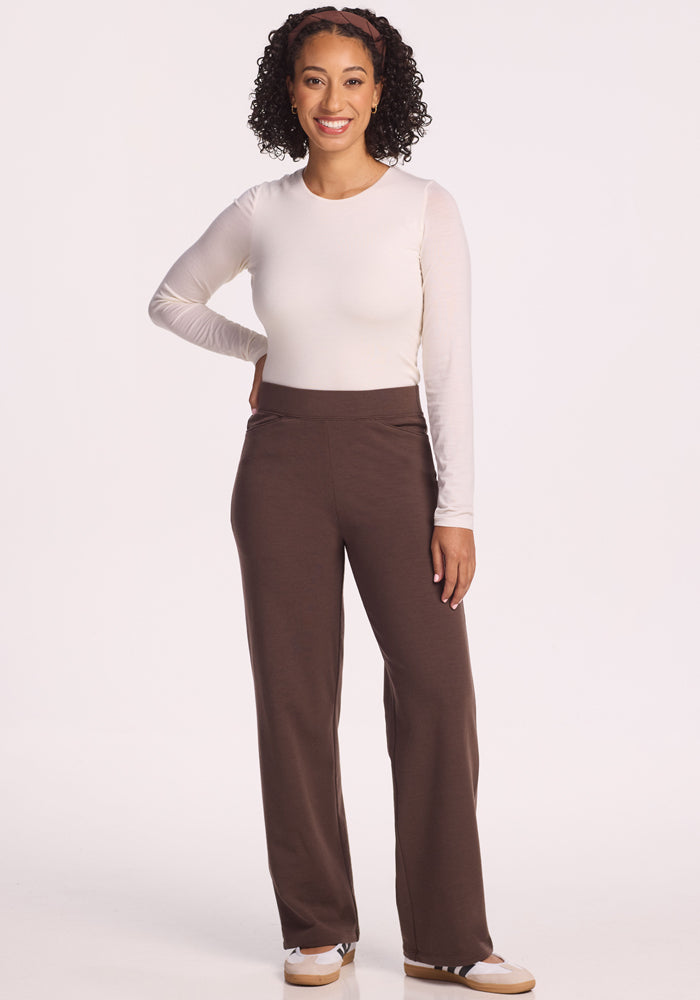 A woman with curly hair smiles while standing against a plain white background, wearing Woolx’s Ellie Wide Leg Pants in Bracken, a white long-sleeve top, and white shoes.