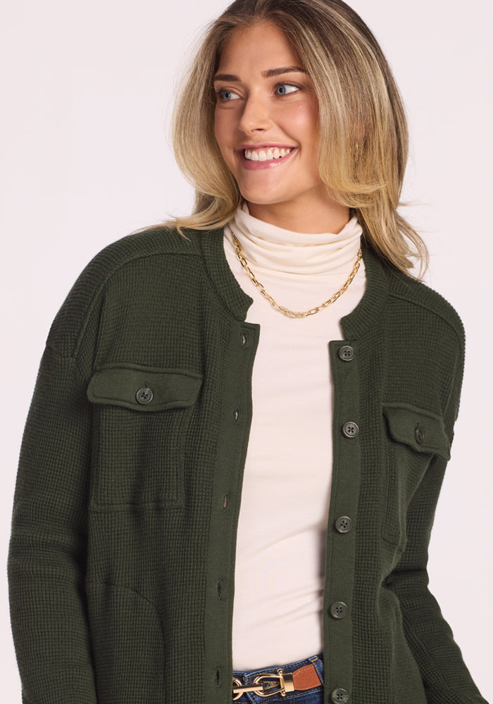 A woman with long blonde hair smiles to the side, wearing the Woolx Oaklynn Waffle Shacket in Driftwood Fern over a white turtleneck and a gold chain necklace. The background is plain and light-colored.