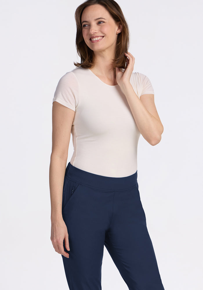 A woman with shoulder-length brown hair smiles while standing against a white background, wearing a light t-shirt and Woolx Nicole Petite Ankle Pant in Deep Navy.