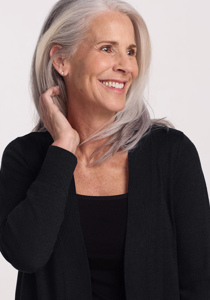 A smiling older woman with long gray hair wears the Woolx Macie Sweater in black over a matching top, looking to her left and gently touching her neck. The background is plain and light-colored.