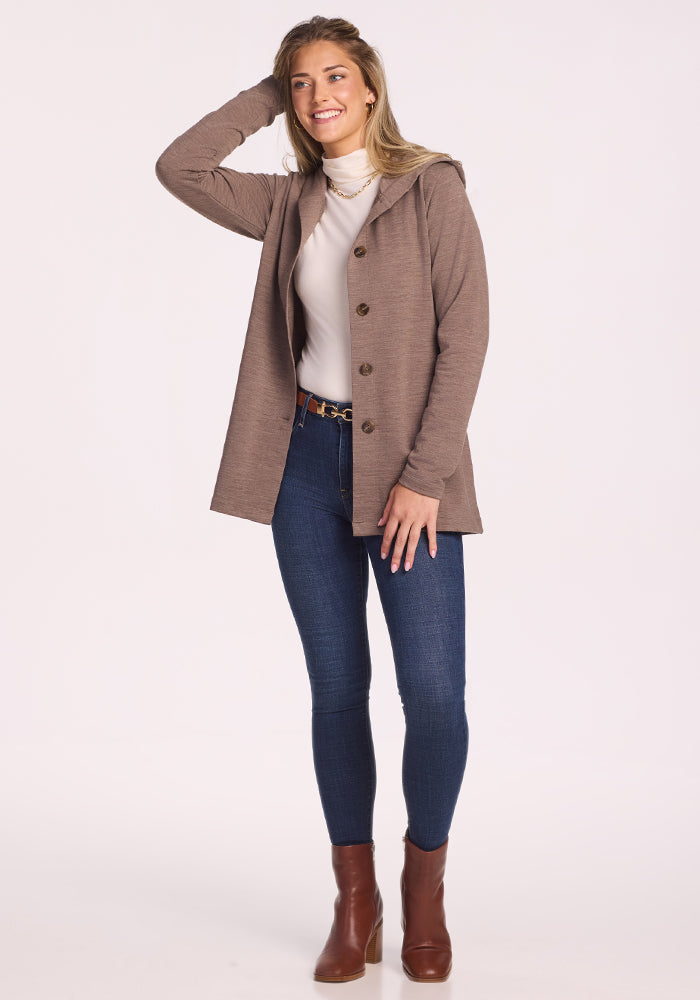 A woman smiles with one hand in her hair, wearing the Woolx Edna Hooded Cardigan in Simply Taupe over a white turtleneck, paired with blue skinny jeans and brown ankle boots, set against a plain light background.