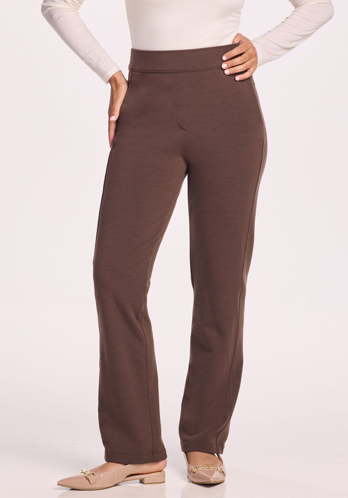 A person wearing Woolx Emerson Straight Leg Pants in Bracken, a long-sleeve cream top, and beige flats with gold chain accents stands with one hand on their hip.