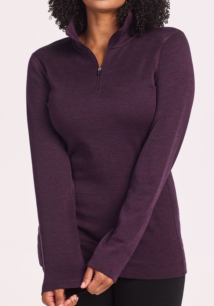 A woman stands with hands clasped, wearing the Woolx Brooke 1/4 Zip in Deep Plum—this Merino wool, long-sleeve pullover with a stand-up collar is perfect as a hiking base layer. Her face is partially out of frame.
