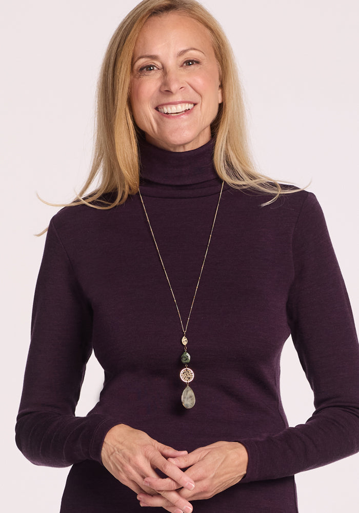 A woman with long blonde hair smiles at the camera, wearing the Woolx Peyton Turtleneck in Deep Plum and a long pendant necklace against a plain light background.