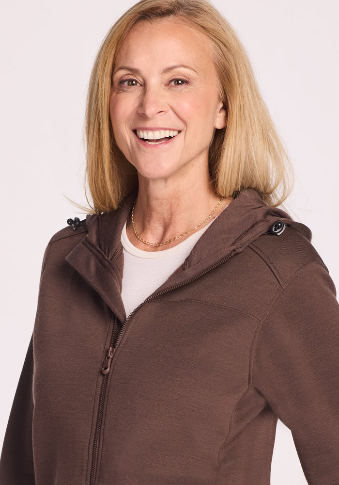 A smiling blonde woman wears the Woolx Cubby Hooded Sweatshirt in Bracken over a white top, standing against a plain light background.