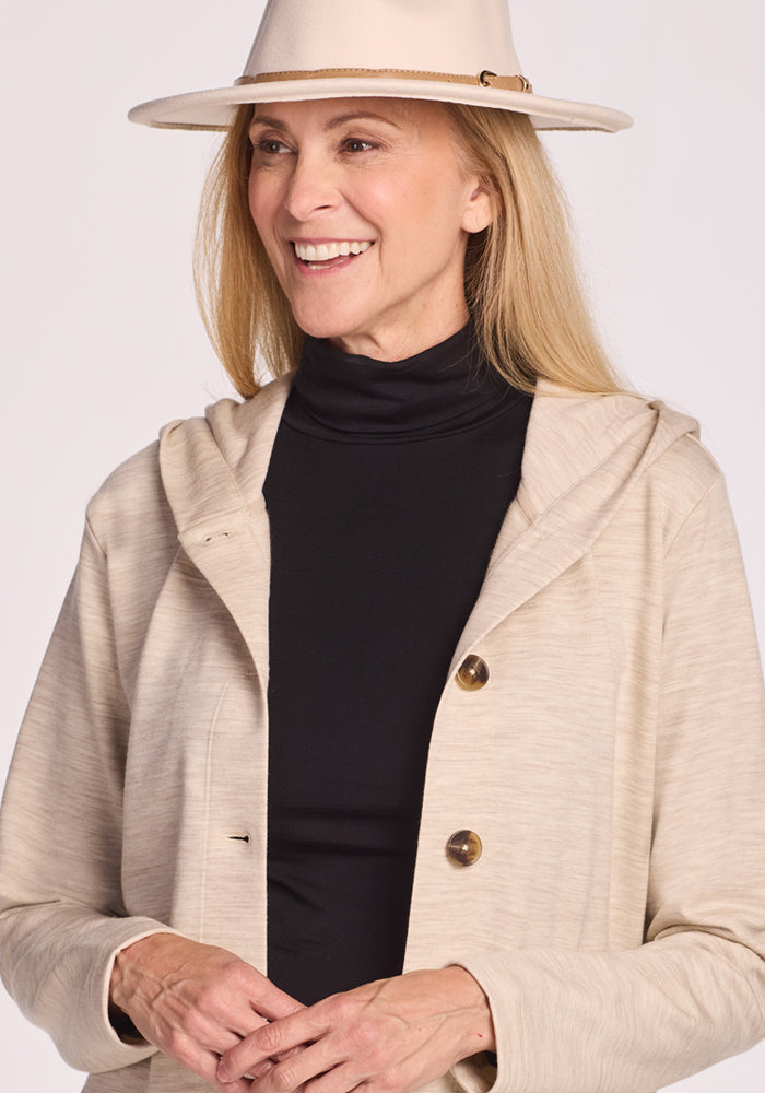 A smiling woman wears the Woolx Edna Hooded Cardigan in Cream Heather over a black turtleneck and beige hat, standing against a light background.