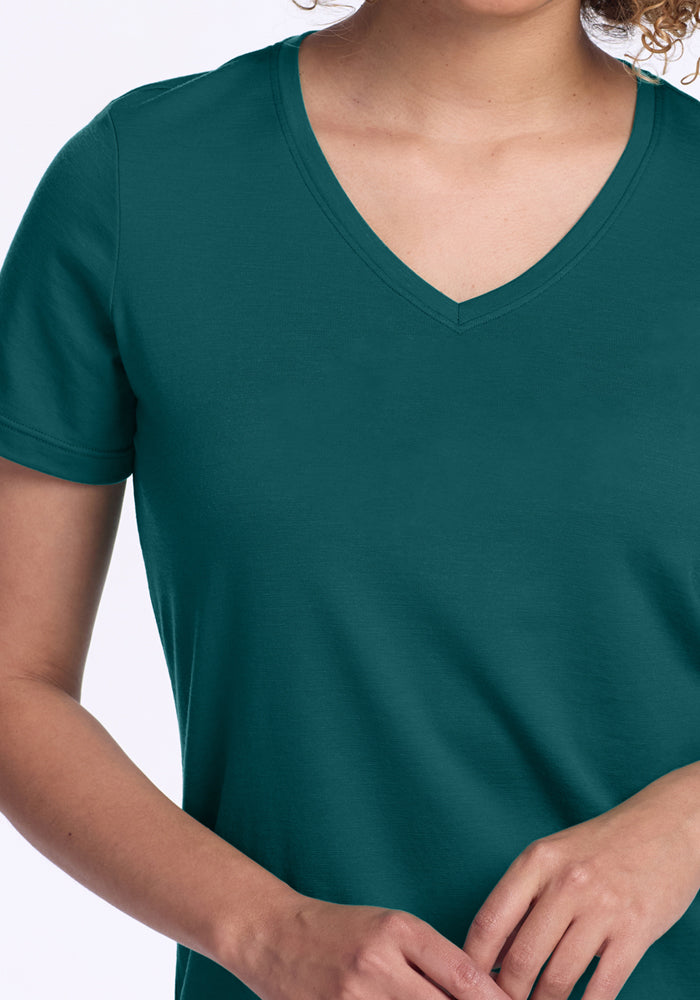 A person wears the Woolx Junie V Neck in Riverbank Green, a relaxed fit Merino wool tee. Against a plain white background, only their upper torso and lower face are visible, with hands gently overlapping near the waist.