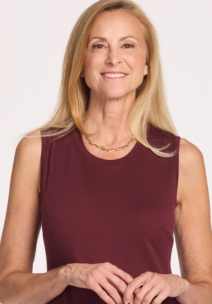 A smiling woman with long blonde hair wears the Woolx Cassie Dress in Toasted Garnet, a sleeveless, temperature-regulating Merino wool tank dress, and a gold chain necklace, standing against a plain light background.