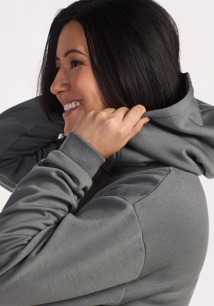 A woman with long dark hair smiles while pulling up the hood of her Woolx Avery Hoodie in Sedona Sage, looking off to the side against a plain light background.