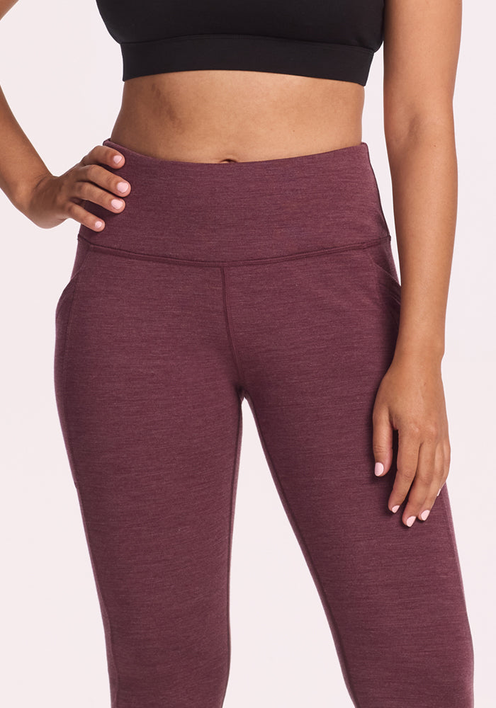 A person stands against a plain light background, wearing Woolx Frost Duralite® Leggings in Grape Harvest Melange with a black crop top, one hand on their hip.