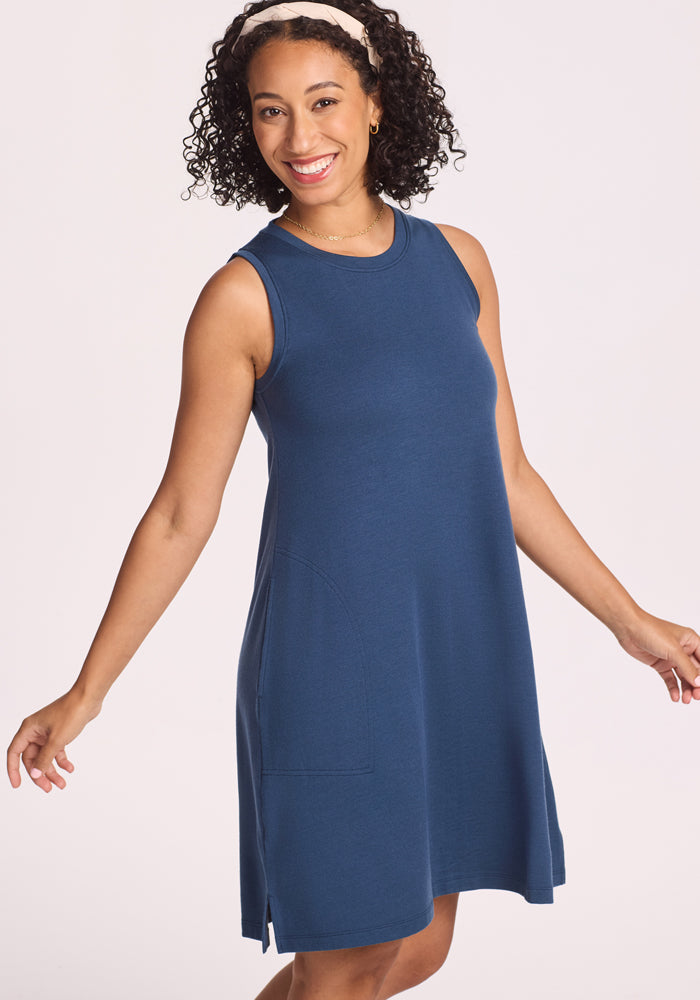 A woman with curly hair wearing a headband smiles while posing in the sleeveless, knee-length Woolx Clara Dress - Starry Night, a blue breathable summer dress with pockets, against a light background.