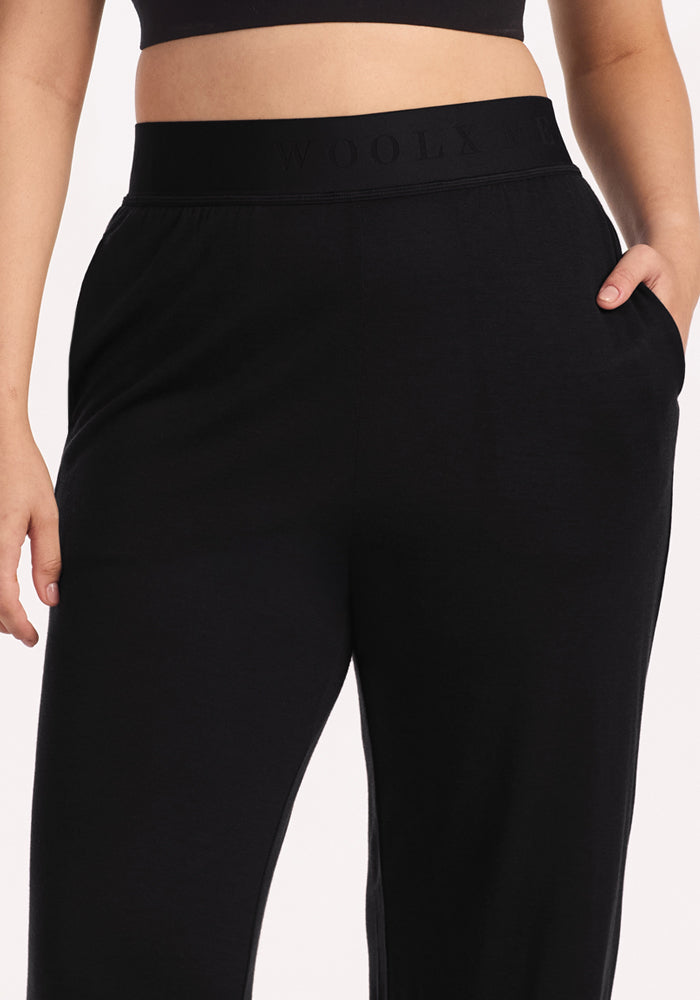 A person wears Woolx Maya Lounge Pants in black, featuring a high-waisted elastic waistband with "WOOLX" branding, paired with a black crop top and hands in the pockets.