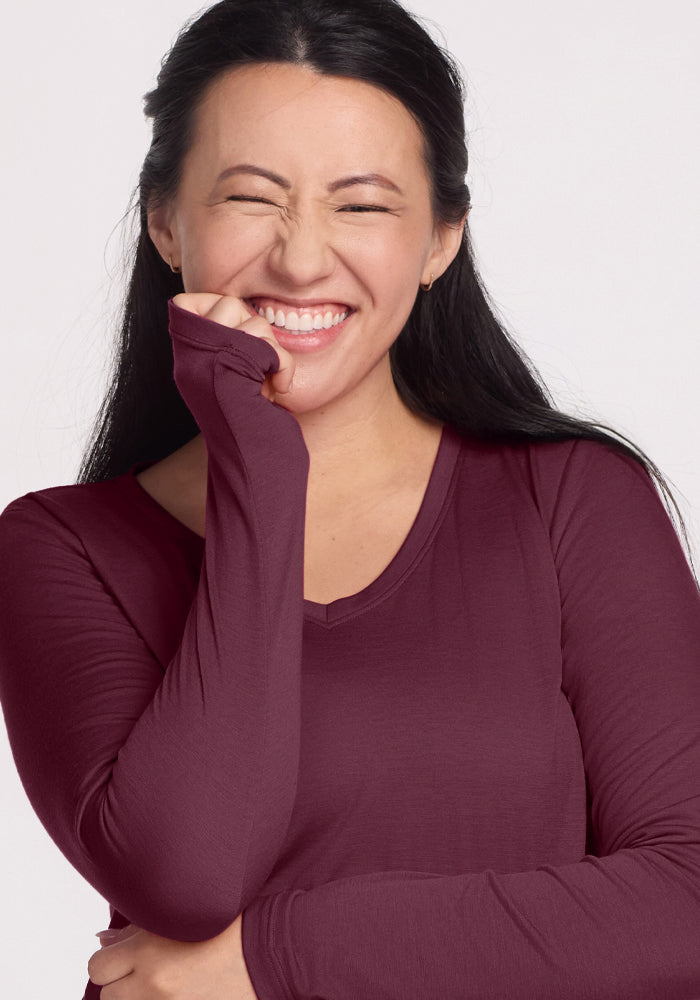 A woman with long dark hair smiles brightly with her eyes closed, playfully covering her mouth while wearing the Woolx Millie Long Sleeve in Grape Harvest, a maroon temperature-regulating Merino wool top.