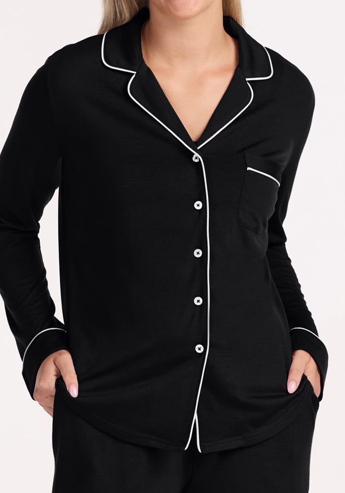 A woman wears the Woolx Harper FeatherTouch® Pajama Set in black, featuring white piping and buttons. Made from moisture-wicking fabric, she stands with hands in pockets against a light background, her face partially out of frame.