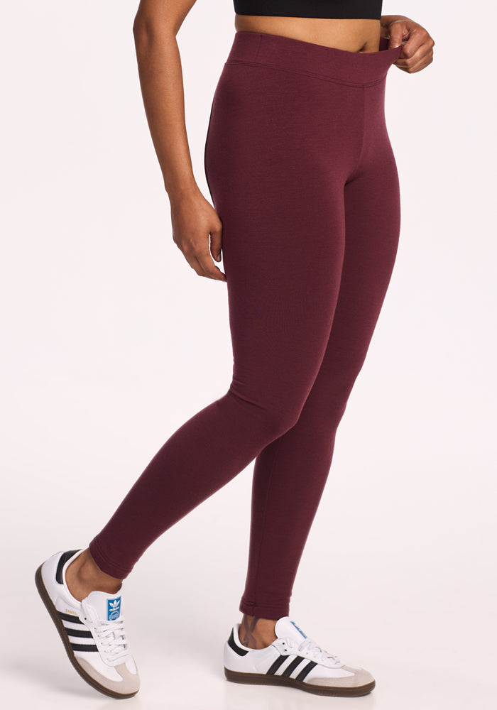 Wearing Woolx Stella Leggings in Toasted Garnet, a black crop top, and white sneakers with black stripes, a person stands against a plain white background, gently pulling at the leggings’ waistband.