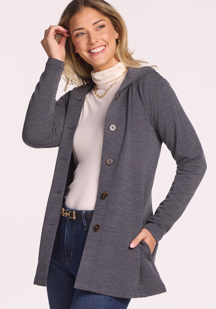 A smiling woman in a light turtleneck, blue jeans, gold necklace, and the Woolx Edna Hooded Cardigan in Cinder Melange poses with one hand in her hair and the other in her pocket—perfect cold-weather style.