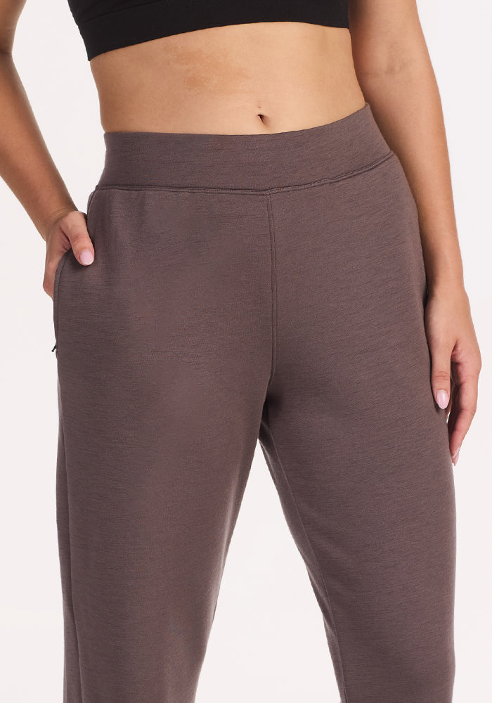 A person wears Woolx Bree Lounge Pants in Plum Truffle, featuring a relaxed fit and high waist, paired with a black crop top, standing with one hand in their pocket against a plain white background.