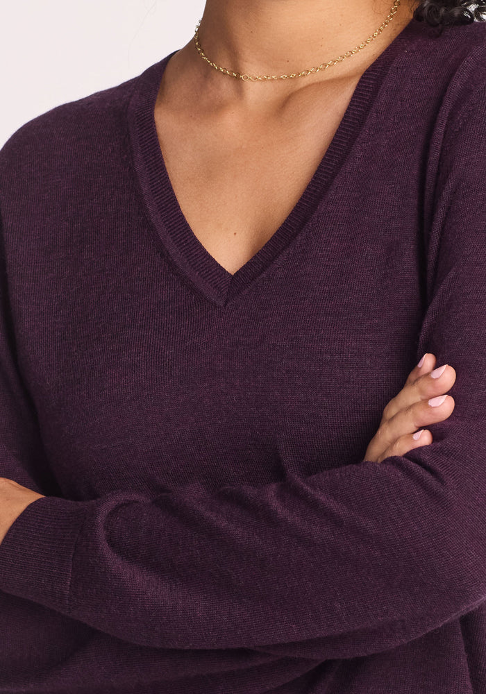A person with arms crossed wears the Woolx Francesca Sweater in Deep Plum and a thin gold chain necklace. The breathable sweater is shown on the upper torso, with the face not visible.