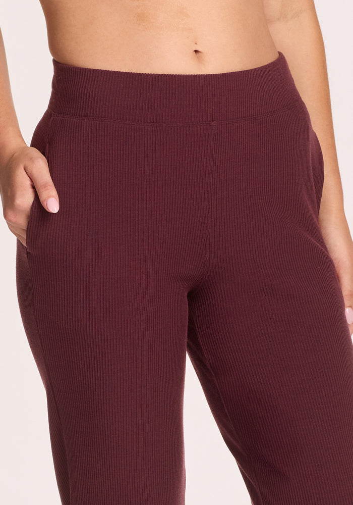 A person wearing Woolx Luca Ribbed Pants in Toasted Garnet, featuring a high waist, wide leg, and ribbed texture, with their left hand in the pocket; the image is cropped from torso to mid-thigh.