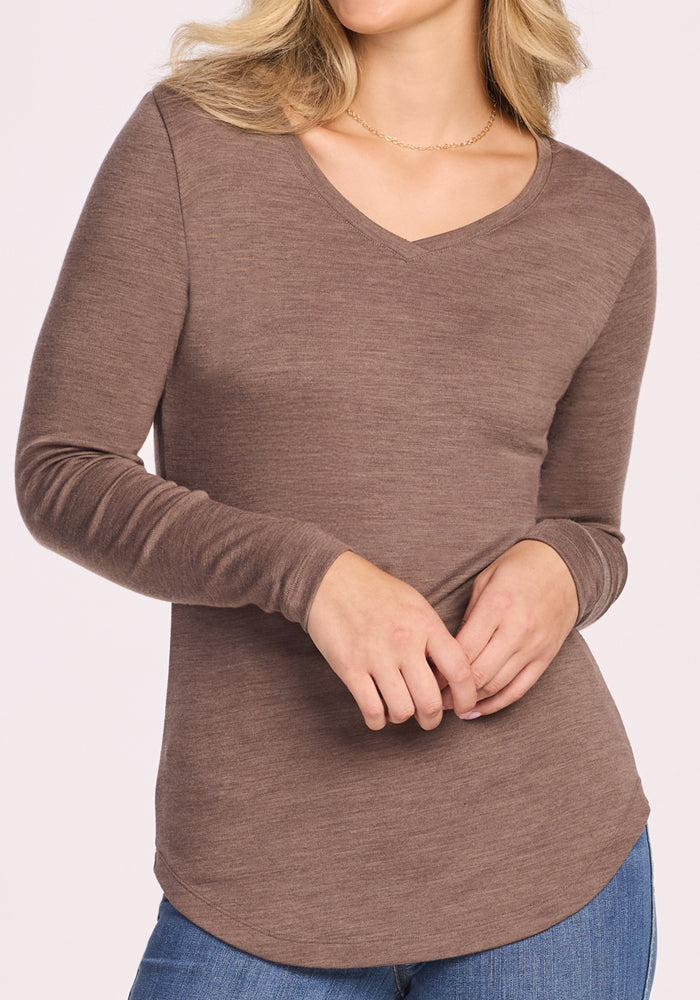 A woman stands with hands clasped, wearing the Woolx Eva Tunic in Simply Taupe and blue jeans. Her face is partially out of frame, showcasing the tunic’s midweight Merino wool warmth and odor-resistant comfort.