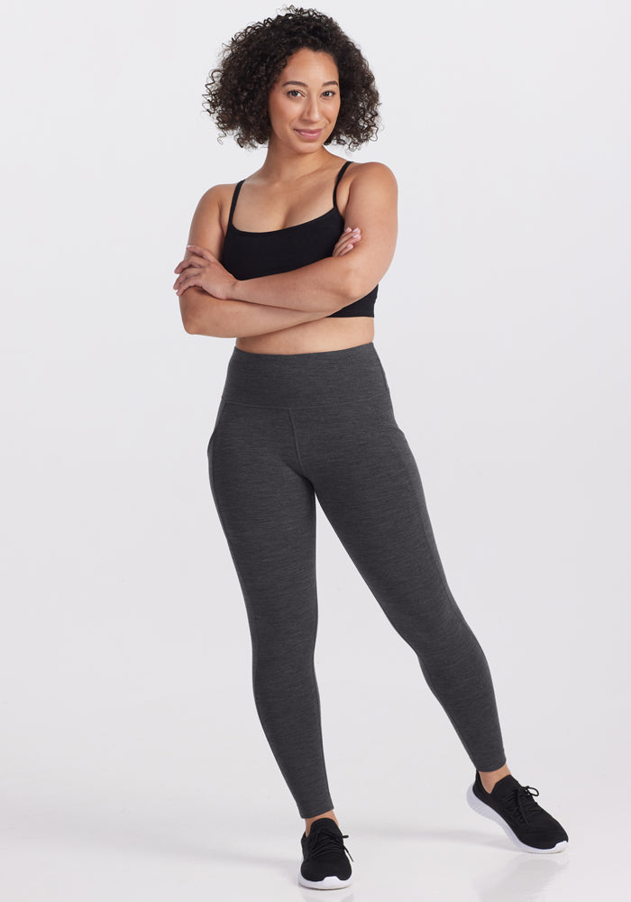 A woman with curly hair, wearing the Woolx Piper Pocket Petite - Cinder Melange leggings, a black sports bra, and black sneakers, stands confidently with arms crossed and smiles against a plain white background.