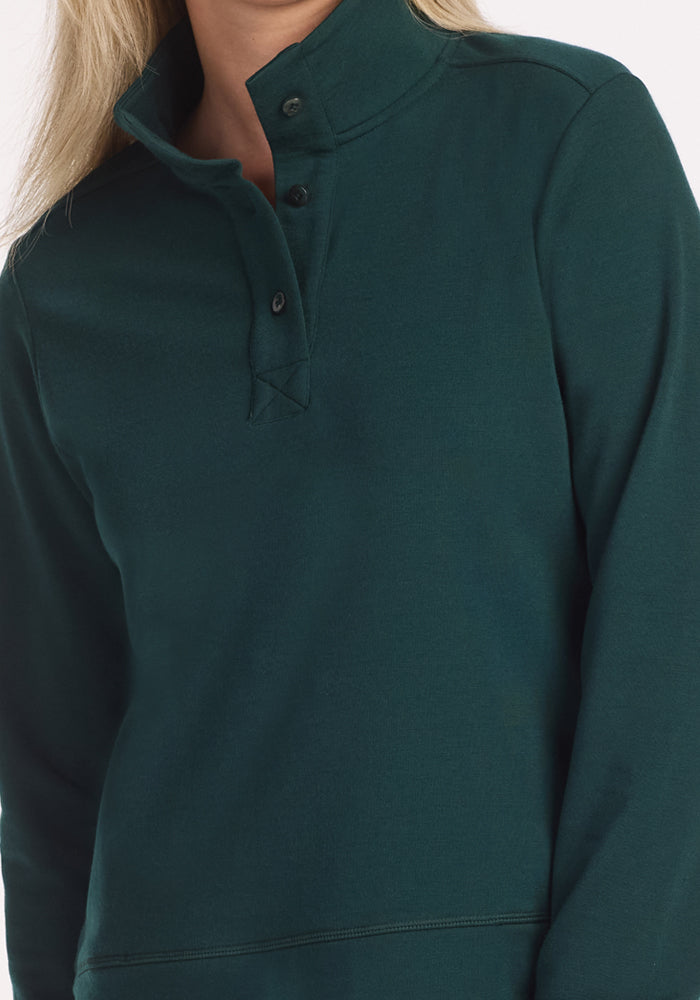 A person wears the Woolx Annie 3 Button Pullover in Deep Timber, a dark green heavyweight sweater with long sleeves and a partially unbuttoned buttoned collar, shown from the upper body only.
