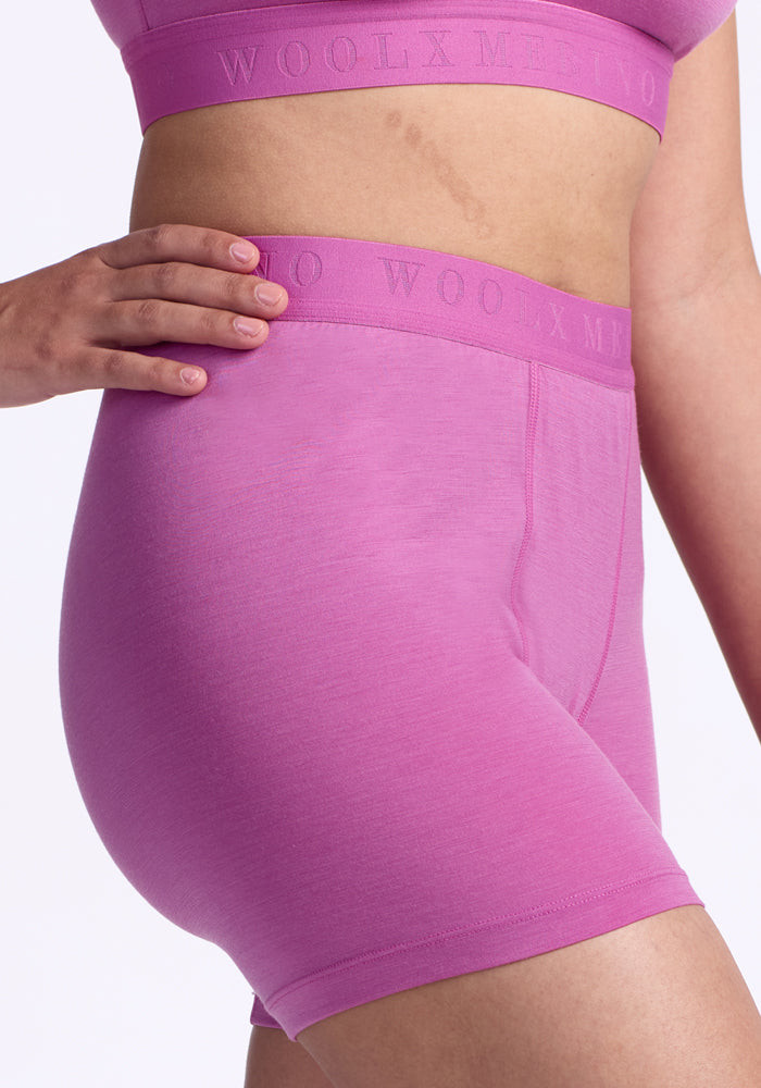 Wearing a matching pink sports bra and Woolx Elliott Boyshort - Wild Blossom with "WOOLX MERINO" on the waistband, they rest their hand on their hip against a plain light background.