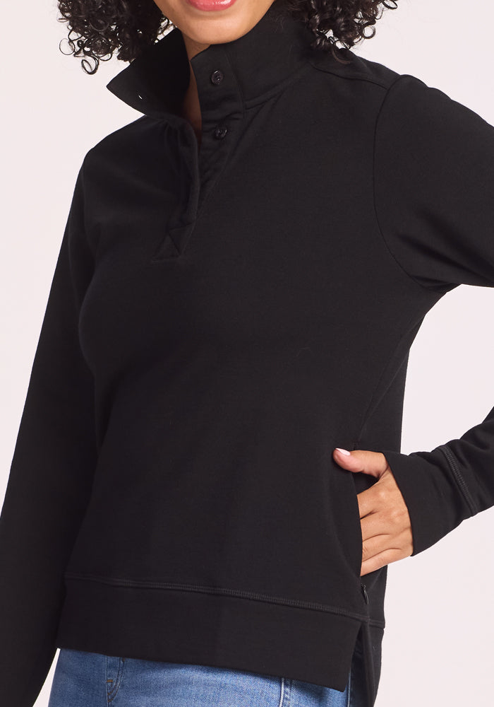 A person wearing the Woolx Annie 3 Button Pullover in black, featuring a high buttoned collar, paired with blue jeans and posing with one hand in their pocket. The face is partially out of frame.