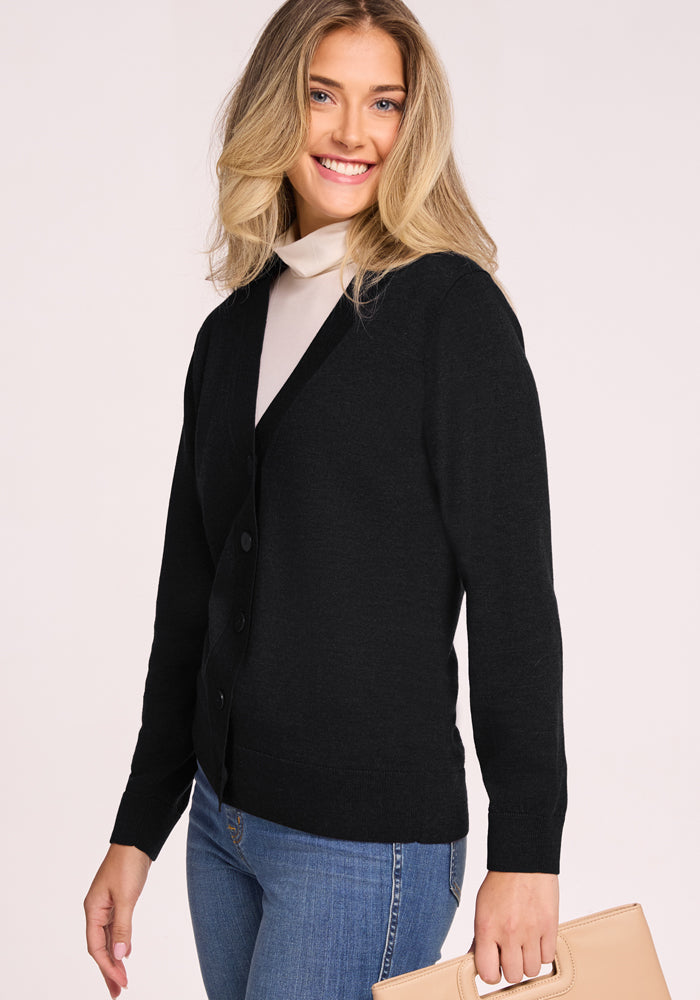 A blonde woman smiles while wearing the Woolx Gia Sweater in black over a white turtleneck and blue jeans, holding a beige handbag against a light background. This lightweight cardigan is both stylish and odor-resistant.