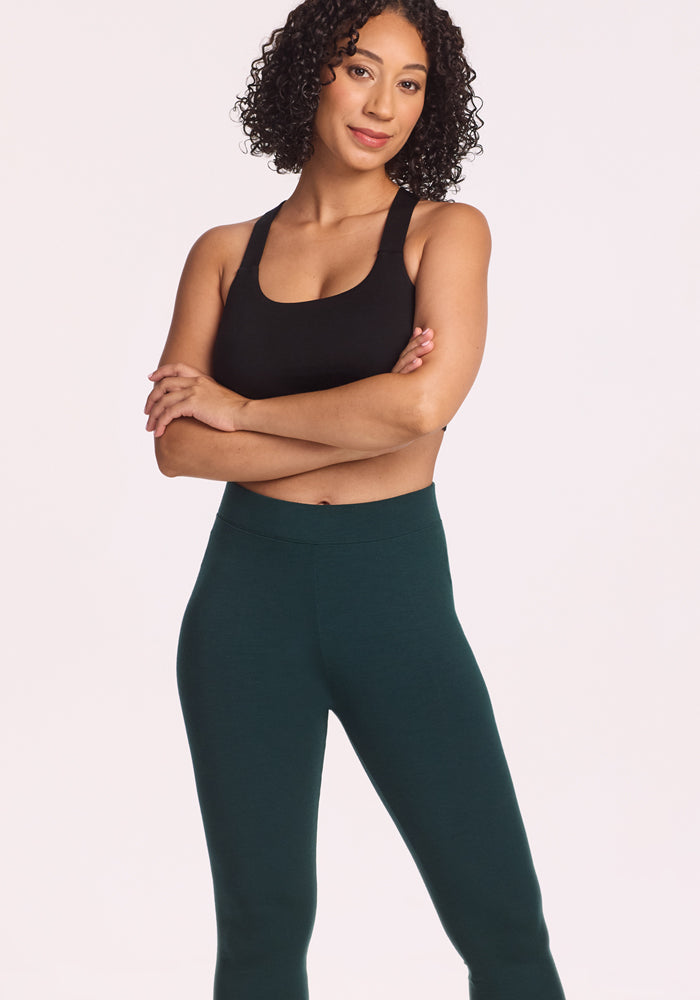 A woman with curly hair stands confidently, arms crossed, wearing a black sports bra and Woolx Stella Leggings in Deep Timber against a plain light background.