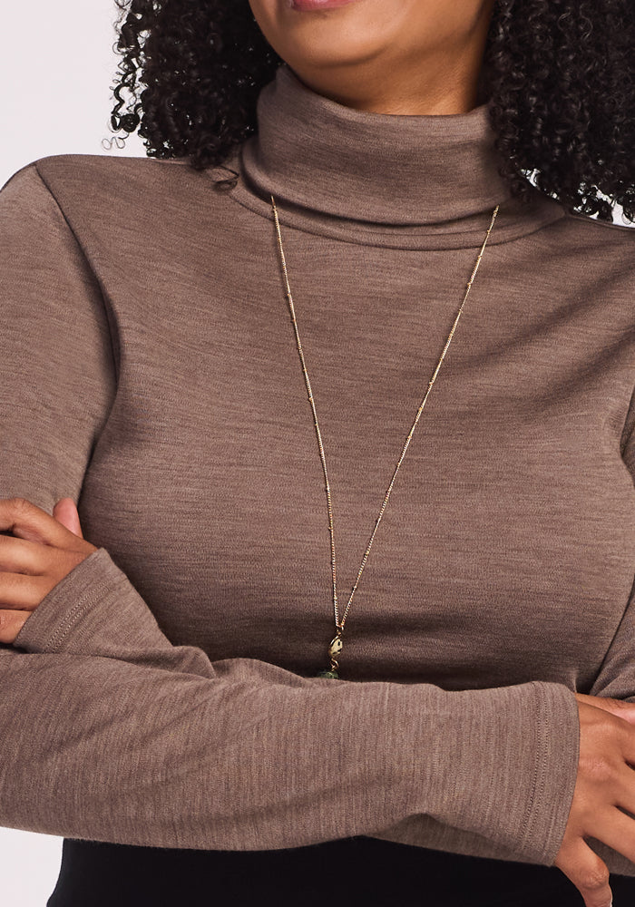 A person with curly hair wearing the Woolx Peyton Turtleneck in Simply Taupe and a long gold necklace, shown from shoulders to mouth with arms crossed.