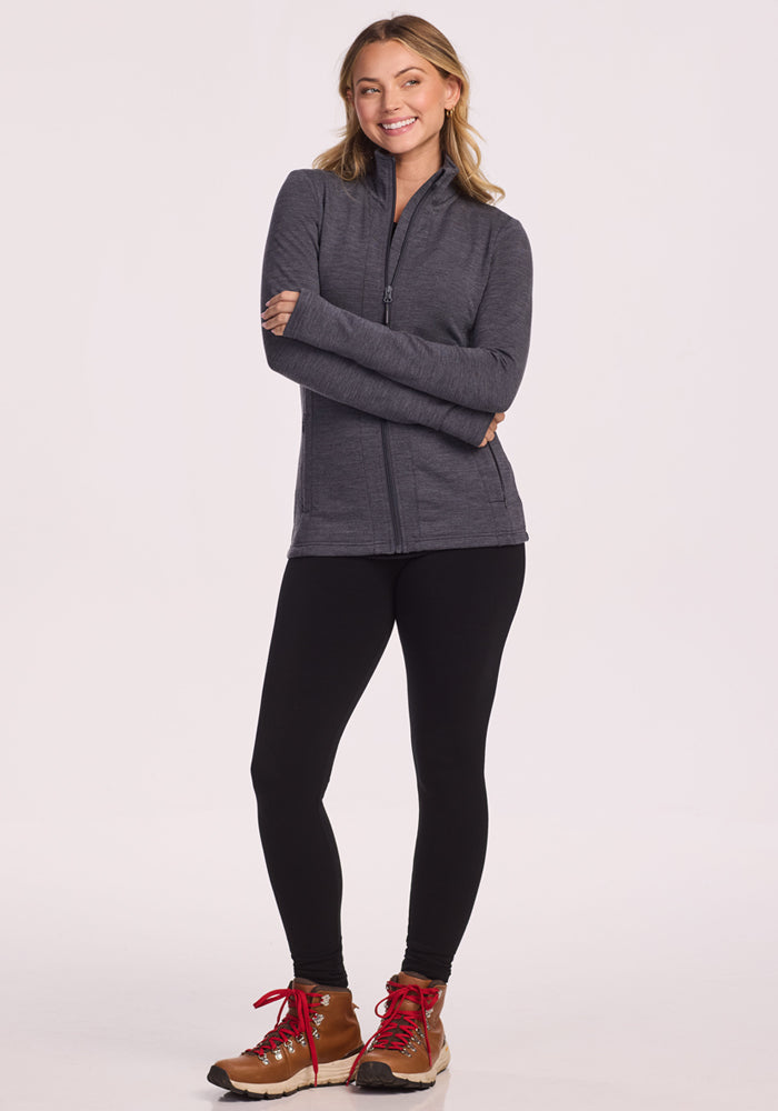 A woman smiles with arms crossed, wearing the Woolx Finley Zip Up Sweatshirt in Cinder Melange, black leggings, and tan hiking boots with red laces against a plain light background.