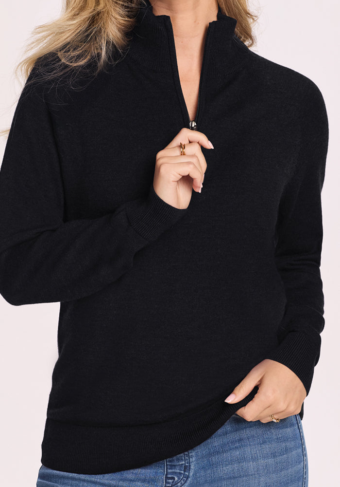 A person is shown from shoulders to hips wearing the Woolx Collins Half Zip Sweater in black and blue jeans, with one hand pulling the zipper and the other resting at the waist.