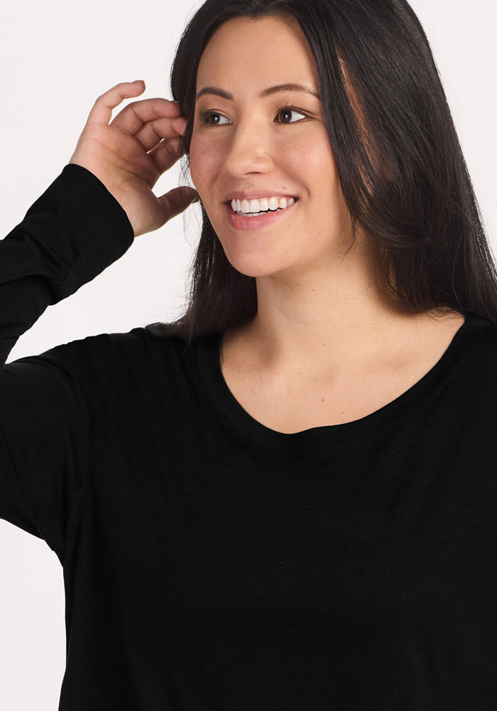 A woman with long dark hair smiles while touching her ear, wearing the Woolx Ashlie Lounge Top in black. The wrinkle-resistant Merino wool travel top is shown against a plain light background as she looks slightly to the side.