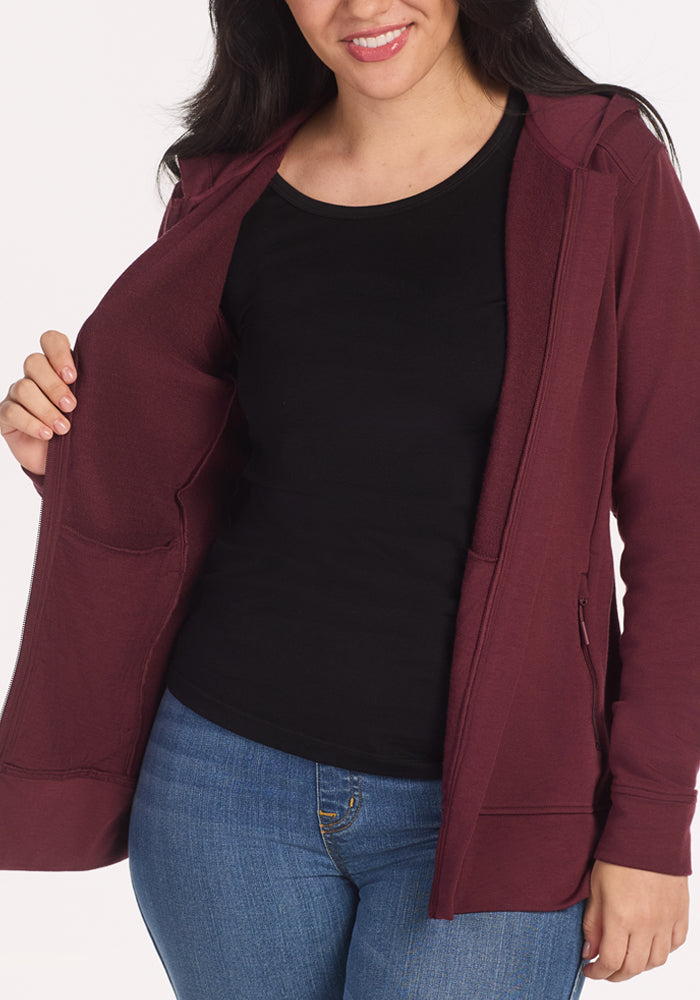 A woman wears the Woolx Darby Zip Up Hoodie in Toasted Garnet over a black shirt and blue jeans, holding one side open to show the convenient inside pocket.