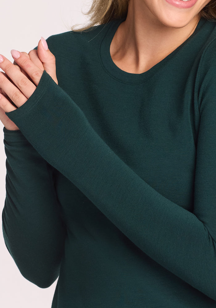 A woman in the Woolx Sadie Baselayer Top - Deep Timber smiles gently, hands raised and fingers touching, showing off the soft Duralite merino fabric and comfortably fitted sleeves.