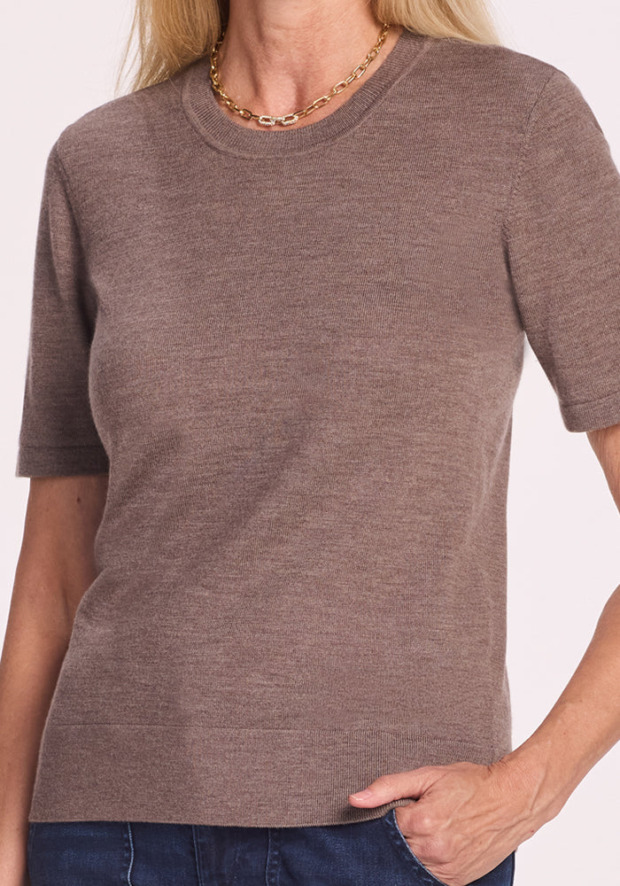 A woman models the Woolx Anna Crew Sweater in Simply Taupe with blue jeans and a gold chain necklace, standing against a plain background with one hand in her pocket. Her face is not visible.