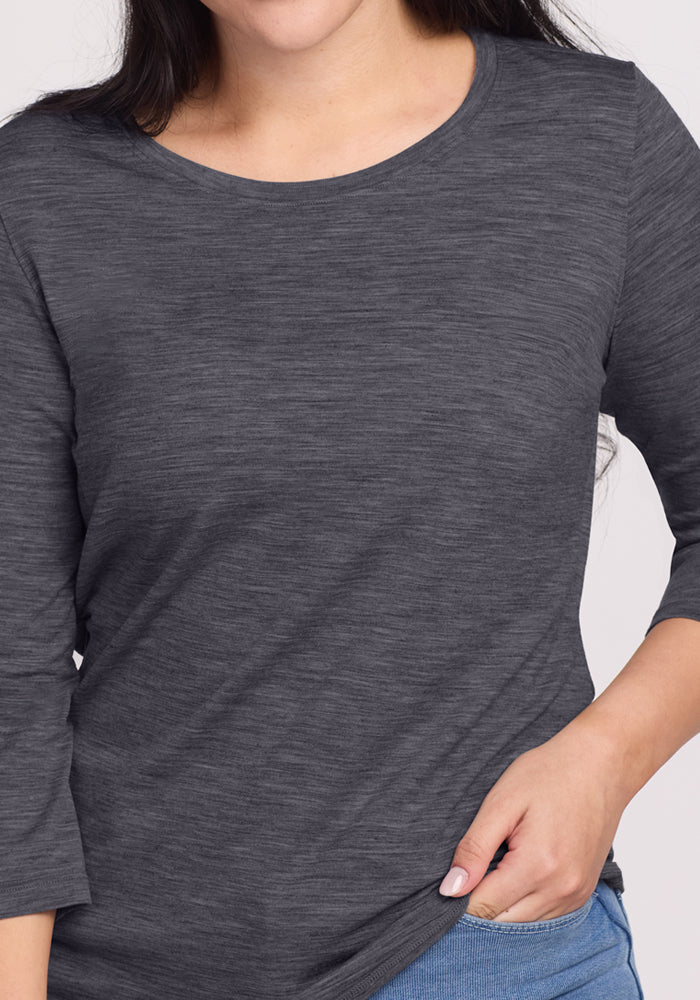 A woman wears the Woolx Jenny 3/4 Sleeve Crew Neck in Cinder Melange with blue jeans, holding the shirt's hem. Her face is partially out of frame.