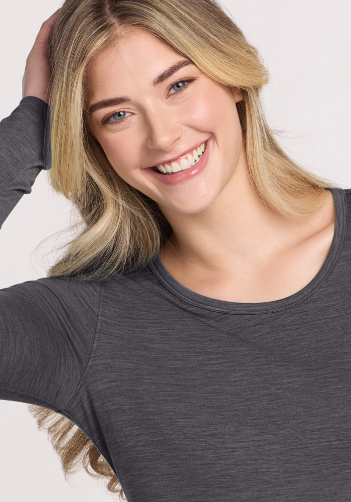 A young woman with long blonde hair smiles brightly, wearing the Woolx Remi Long Sleeve T-Shirt in Cinder Melange. She raises one hand to rest on the back of her head, set against a light, plain background.