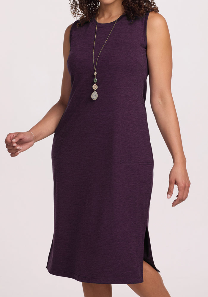 A woman in the Woolx Cassie Dress in Deep Plum, a sleeveless knee-length Merino wool tank with side slits, accessorized with a long pendant necklace, stands against a plain light background.
