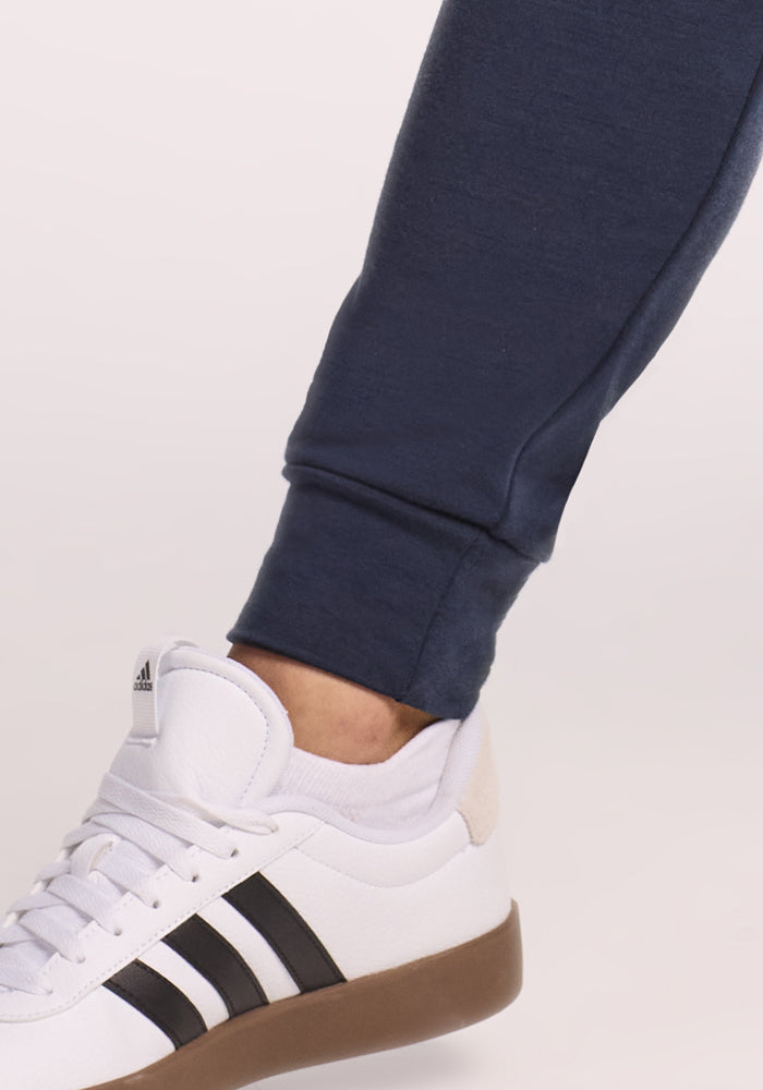 Close-up of a person wearing Woolx Willa Joggers in Deep Navy paired with white Adidas sneakers with black stripes and a light brown sole, standing on a light-colored surface.