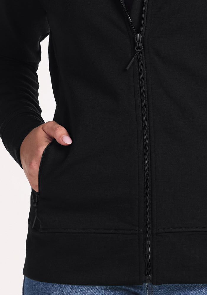 A person wears the Woolx Darby Zip Up Hoodie in black, made from soft Duralite fabric, with their left hand in the pocket. Light blue jeans are partially visible at the bottom.