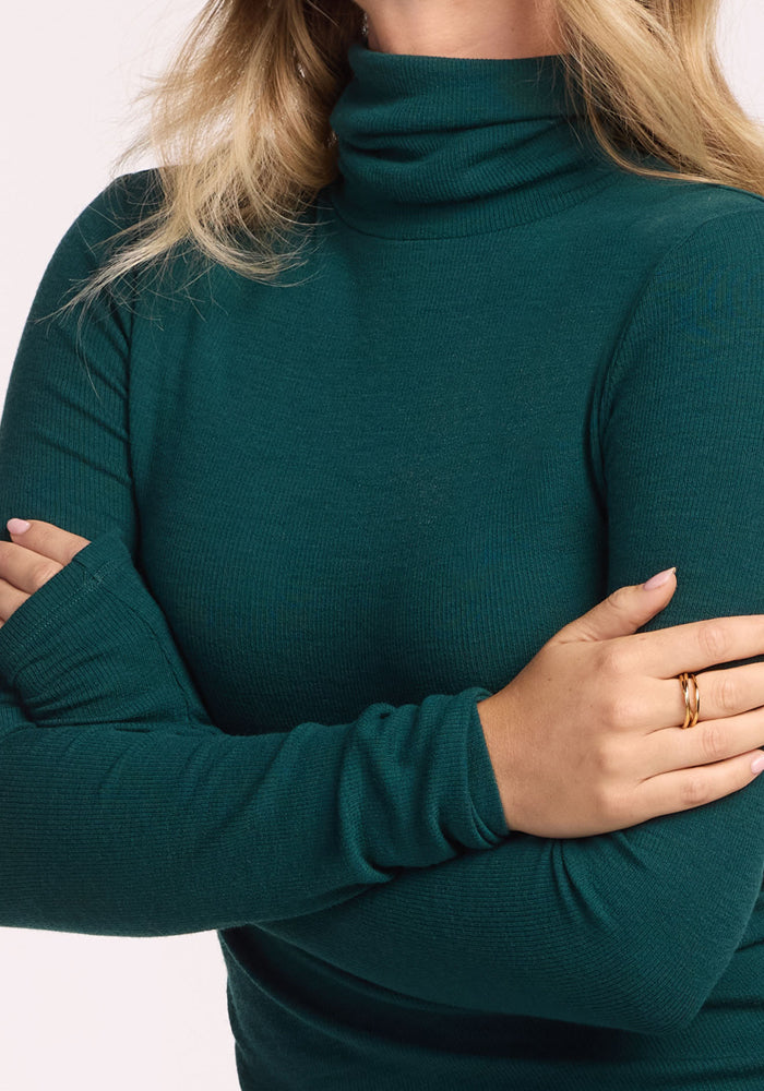 A person with blonde hair wears the Woolx Sage Ribbed Turtleneck in Riverbank Green and gold rings, standing with arms crossed against a plain background.