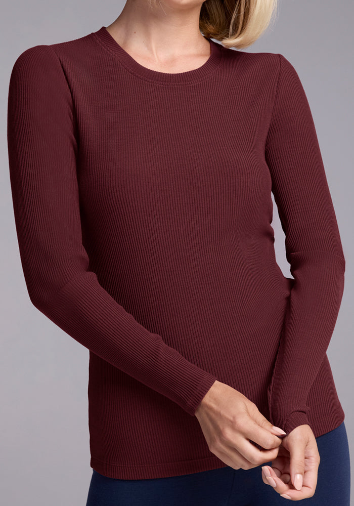 A woman adjusts the sleeve cuff of the Woolx Hadley Ribbed Crew in Toasted Garnet, a lightweight merino wool long-sleeve top perfect for travel. She stands against a gray background, her face partially out of frame.