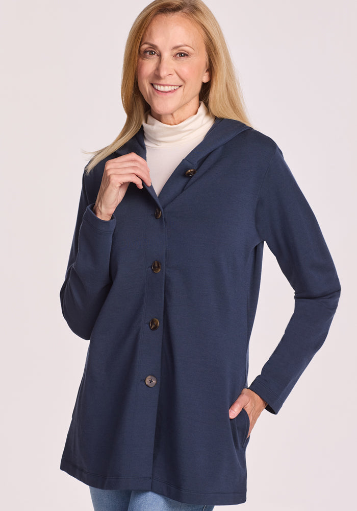 A smiling woman with long blonde hair wears the Woolx Edna Hooded Cardigan in Deep Navy over a white turtleneck, one hand near the collar and the other in her pocket, standing against a plain light background.