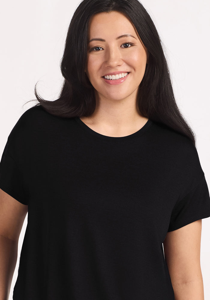 A woman with long dark hair, wearing the Woolx Billie Lounge Tee in black, smiles at the camera against a plain light background.
