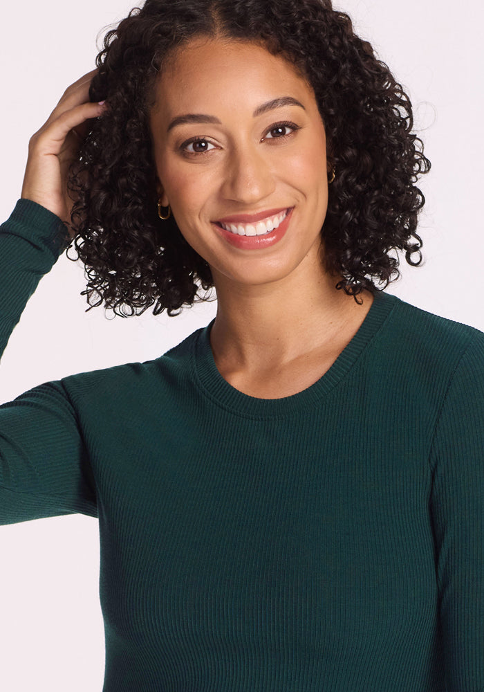 A woman with curly dark hair, wearing the Woolx Hadley Ribbed Crew in Deep Timber, smiles warmly at the camera and touches her hair with one hand. The background is plain white.