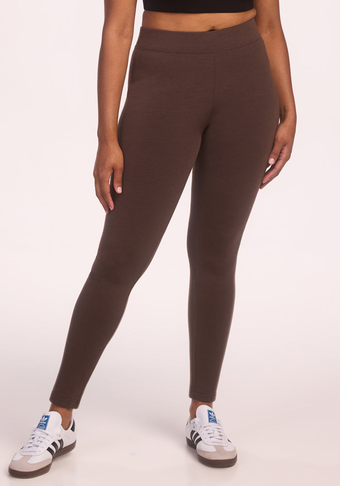 Wearing Woolx's Stella Tall - Bracken leggings, a black cropped top, and white sneakers with blue and gray accents, a person stands against a plain light background. Only the body from shoulders down is visible.