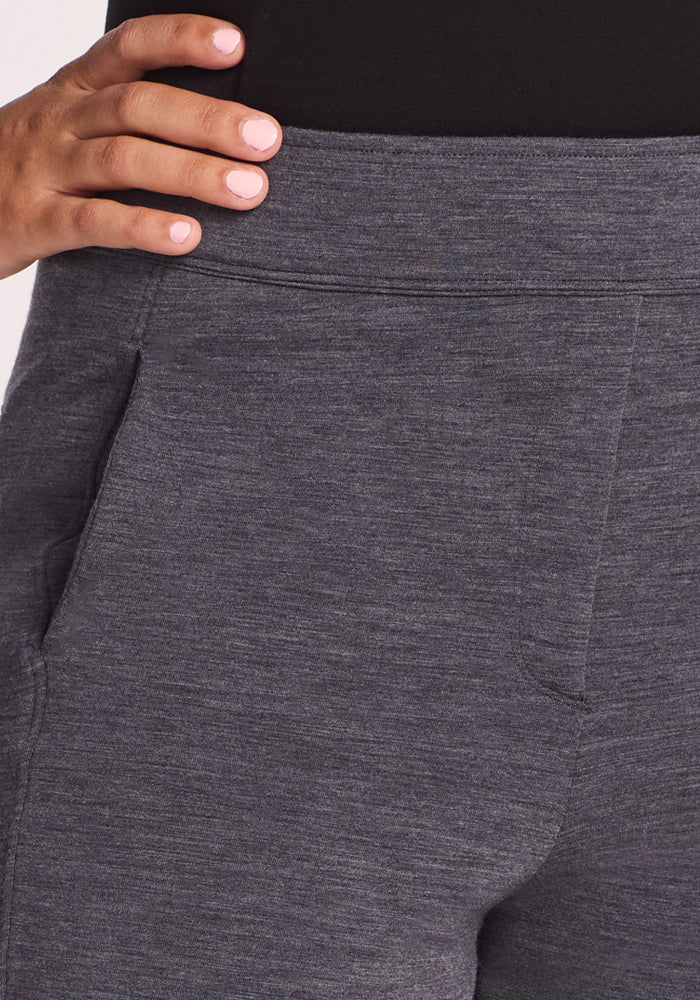 A close-up shows a person wearing Woolx Emerson Straight Leg Pants in Cinder Melange and a black top, with their left hand on their hip. The pants feature visible pockets and a heathered texture.
